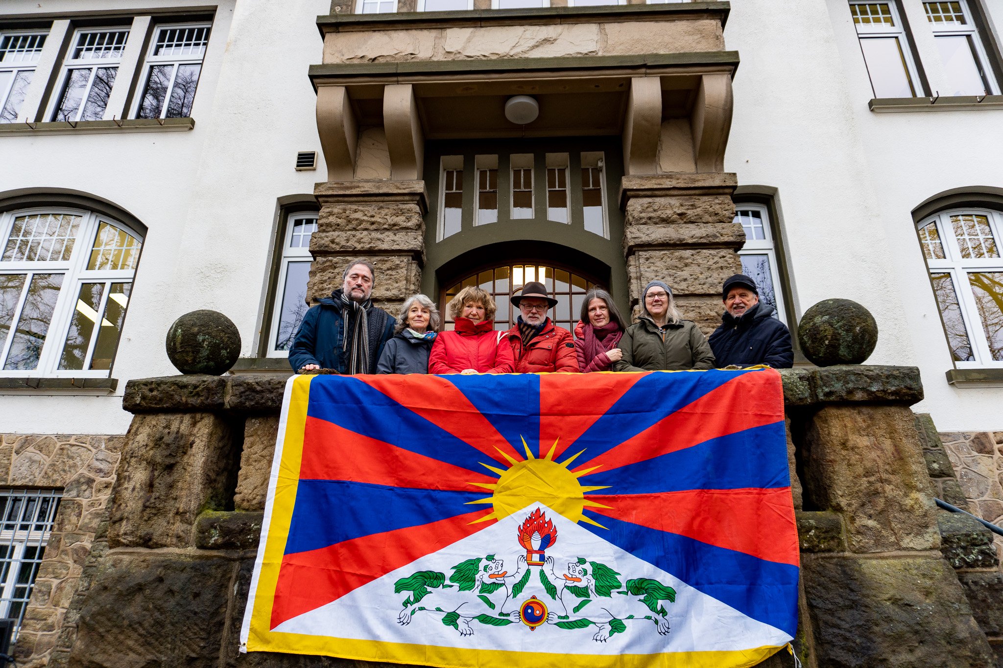 Menschen stehen auf einem Balkon, an dem eine bunte Fahne hängt